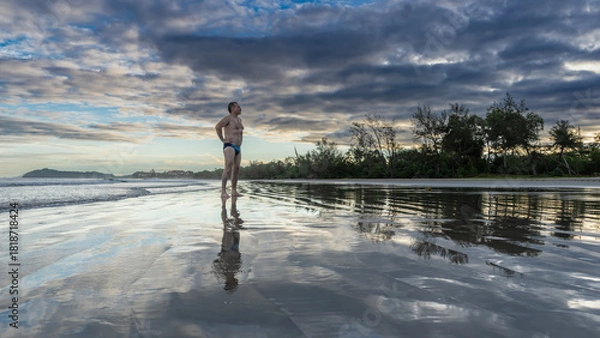 Fototapeta A man stands on the ocean shore at dawn, looking into the distance. Hands on the waist. Waves are spreading across the beach. Reflection on wet, smooth sand.The clouds are highlighted in pink and gold