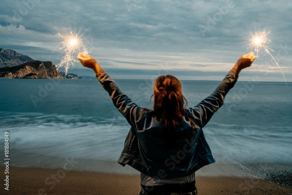 Fototapeta Back view of young happy smiling woman holding sparklernear the sea . Christmas, New Year, concept.