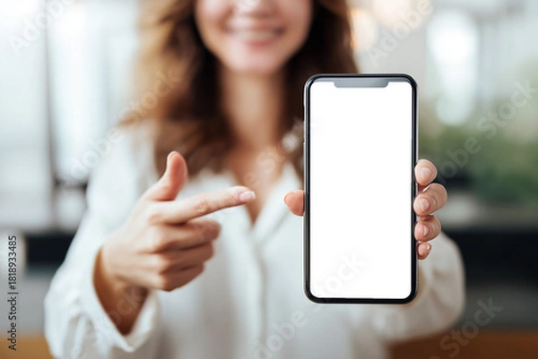 Obraz A smiling woman in a white top holds out a smartphone with a blank black screen and points at it, isolated as a PNG with a transparent background.