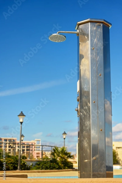 Obraz High outdoor metal shower with sun glare, reflections, scratches, and dried water stains. Selective focus on the foreground; blurred background with lampposts and hotel buildings