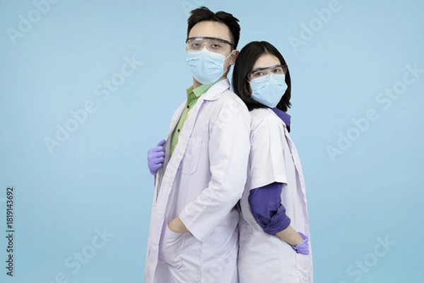 Obraz Confident Asian medical professionals in lab coats and safety gear, standing back-to-back, symbolizing strong teamwork and healthcare in blue studio background