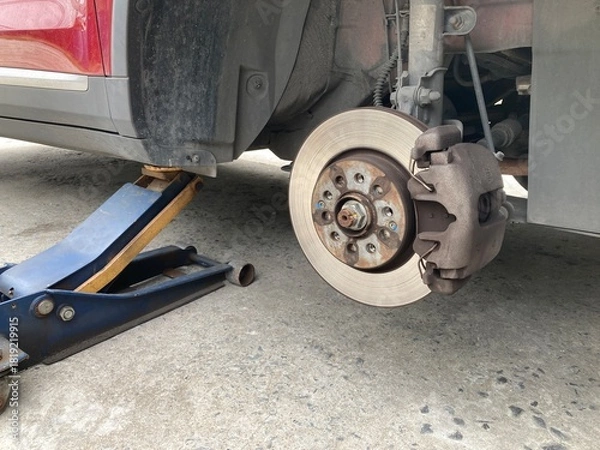 Fototapeta Low-angle view of a red car lifted by a hydraulic jack on a concrete floor. The wheel is removed, showing the brake disc and caliper assembly during a DIY repair or maintenance process.