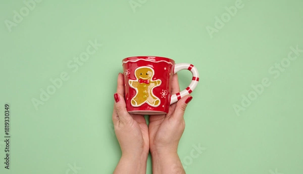 Obraz Two female hands holding a red Christmas mug with a gingerbread man on a green background.