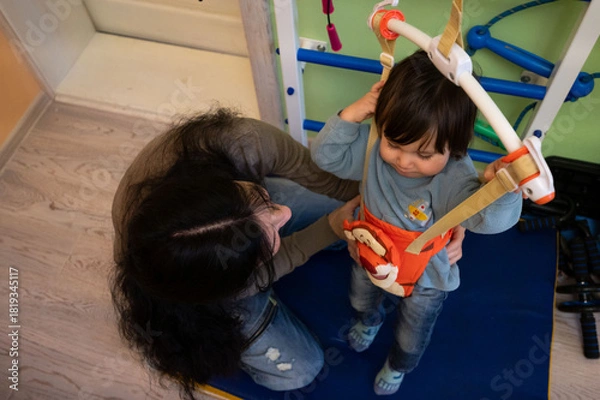 Fototapeta Child engaging in playful movement with support harness, while caregiver encourages exploration during activity session in a cheerful indoor play area