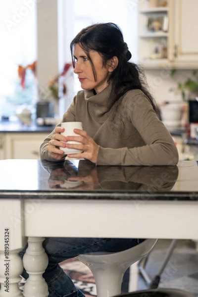 Fototapeta Contemplative woman holding a warm beverage while seated at a kitchen table in a cozy, well-lit home setting at midday