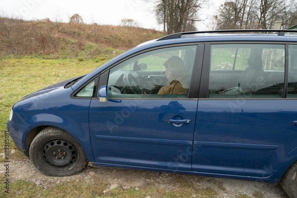 Fototapeta Family outing to the countryside in early spring with a blue minivan parked on a grassy meadow while passengers enjoy the fresh air and scenic views