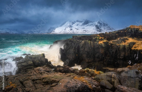 Fototapeta Wave impact on a rocky coastline with turquoise sea and snowy mountains in Lofoten Islands, Norway. Dynamic coastal landscape with cliffs, winter colors and rough water. Dramatic scenery with rocks