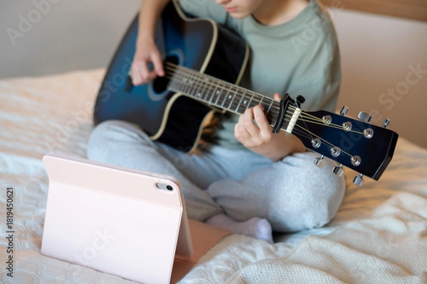 Fototapeta A teenage girl sits with a guitar, learning to play using a tablet program, illustrating online music learning, digital technology, and engaging leisure activities at home