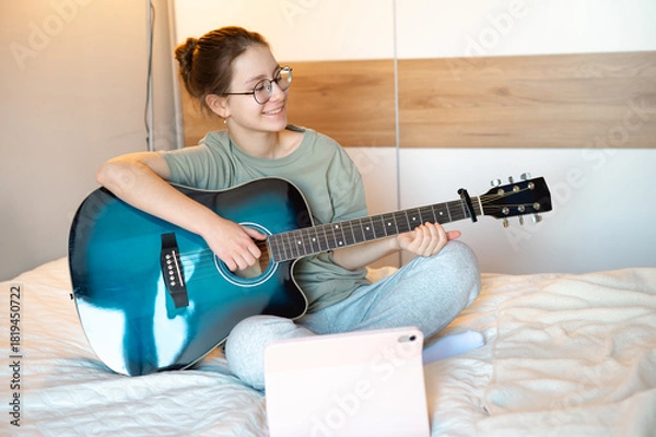 Fototapeta A teenage girl plays the guitar, showing it to someone online while smiling, illustrating joy of music, digital interaction, and engaging leisure activities at home