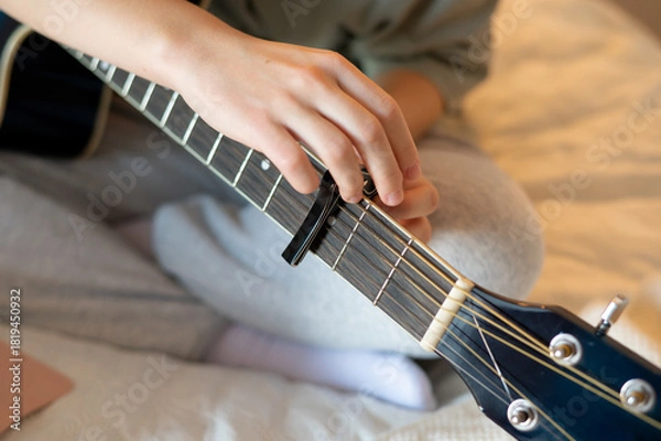 Fototapeta Close-up of a girl’s hands attaching a capo on her guitar’s neck and playing, showing hands-on practice, precise movements, and engagement with music in a home environment
