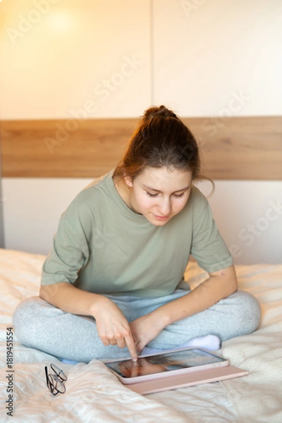 Fototapeta A teenage girl sits on her bed in comfortable home clothes, focused on a tablet, illustrating everyday leisure, digital technology, and a cozy home atmosphere in a relaxed setting