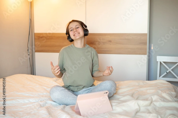 Fototapeta A teenage girl wearing headphones sits on her bed in casual home clothes, happily watching or listening to a tablet, illustrating digital leisure and a cozy, joyful home atmosphere