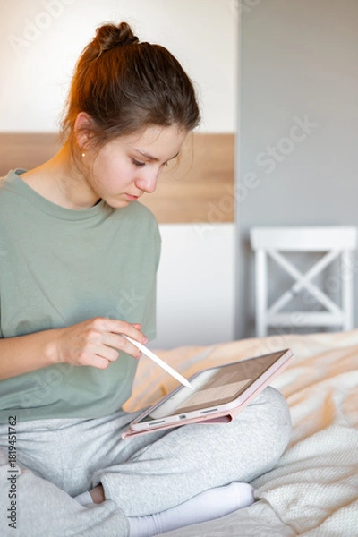 Fototapeta A teenage girl sits on her bed in comfortable home clothes, focused on a tablet, illustrating everyday leisure, digital technology, and a cozy home atmosphere in a relaxed setting