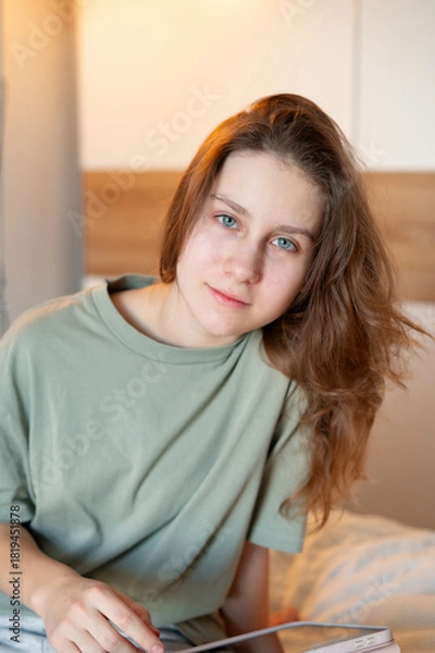 Fototapeta A teenage girl sits on her bed in comfortable home clothes, focused on a tablet, illustrating everyday leisure, digital technology, and a cozy home atmosphere in a relaxed setting