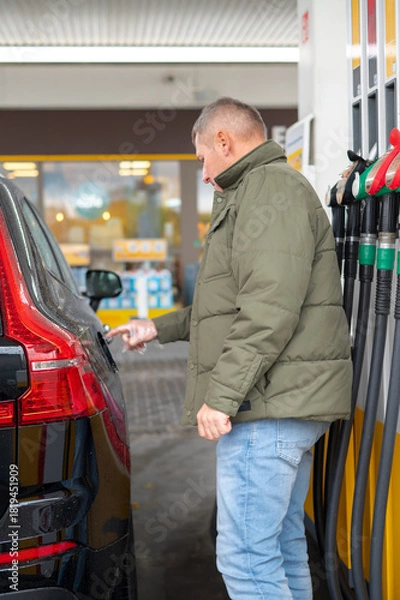 Fototapeta A young man opens a car’s fuel tank flap at a gas station, preparing to refuel, showing everyday refueling, human interaction with vehicles, and the development of urban infrastructure