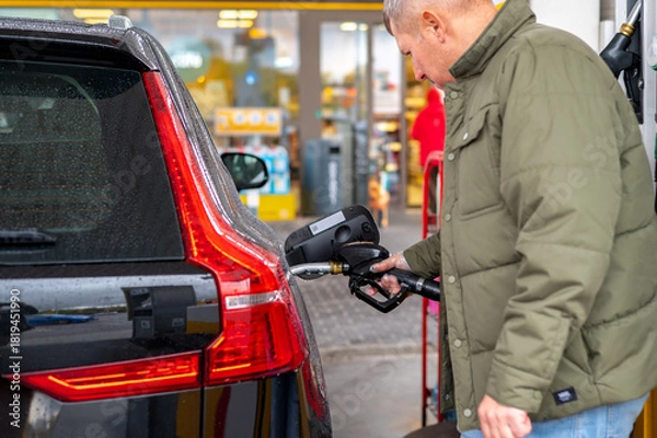 Fototapeta A man inserts a fuel nozzle into a car’s tank at a gas station, showing the refueling process, human interaction with vehicles, and the development of urban infrastructure for automobiles