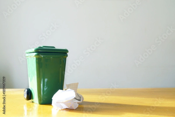 Obraz A small green waste bin with crumpled paper on a wooden table stands in natural light. Highlighting sustainable living, eco habits, minimal waste, recycling culture, and environmental awareness. 