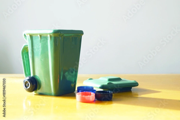 Obraz Green small waste bin with colorful bottle caps arranged as sorting demonstration. Concepts of plastic garbage separation, micro-plastic pollution awareness, and waste segregation education.