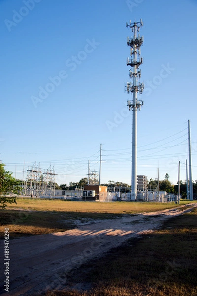 Fototapeta Cell Tower Antenna Mast Rises Against Clear Blue Sky With Wires and Infrastructure. Tall telecom tower with numerous antennas stands against a vivid blue sky; wires and cables run at its base, highlig