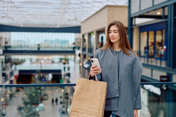 Obraz Young woman buying items on smartphone, holding a shopping bag in a modern environment. Concept of mobile commerce and retail