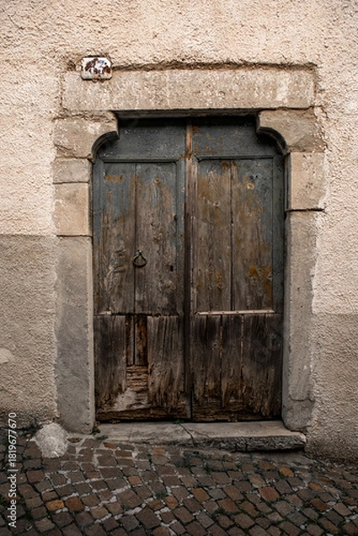 Fototapeta Old wooden door showing decay, rustic texture, and a simple metal knocker, set in a historic stone doorway