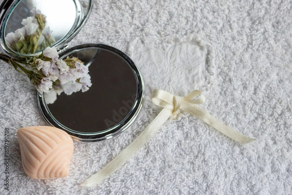 Obraz Compact mirror reflecting delicate flowers lying on a plush white towel with a decorative soap bar and a cream ribbon, symbolizing beauty, self care, and a calming spa atmosphere