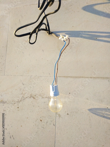 Obraz The light bulb hangs against a light concrete wall, illuminated by sunlight. Subtle shadows and traces of time are visible on the surface, creating an atmosphere of simplicity and everyday life.