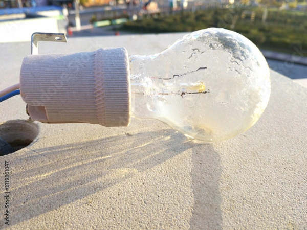 Obraz The light bulb hangs against a light concrete wall, illuminated by sunlight. Subtle shadows and traces of time are visible on the surface, creating an atmosphere of simplicity and everyday life.