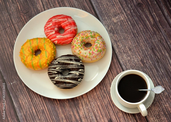 Obraz Four fresh doughnuts with colorful glaze and toppings and a cup of hot black coffee on a wooden table.
