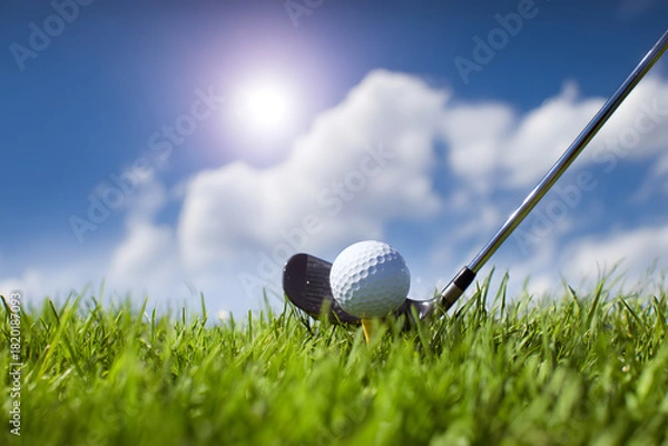Fototapeta Close-up of a golf ball poised for a swing next to a golf club on a lush green fairway, bathed in sunlight with a blue sky and fluffy clouds in the background. Sports concept