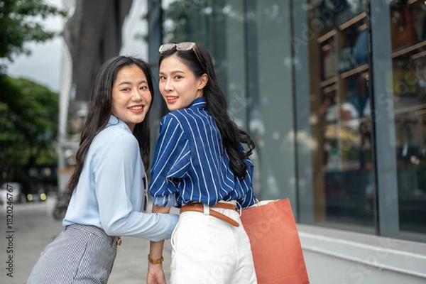 Obraz Asian woman friends holding shopping bag linking arm turning looking back while walking outside mall