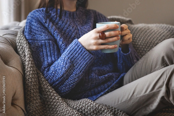 Obraz Female hands holding blue mug while sitting in oversized knitted sweater and warm blanket. Cozy autumn or winter atmosphere indoors with comfort