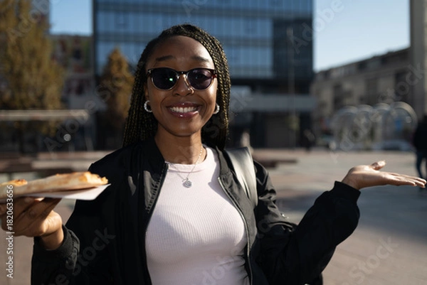 Obraz Beautiful black woman with sunglasses eating a slice of pizza on the city street on a sunny day