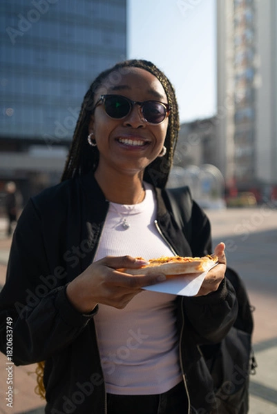 Fototapeta Beautiful black woman with sunglasses eating a slice of pizza on the city street on a sunny day