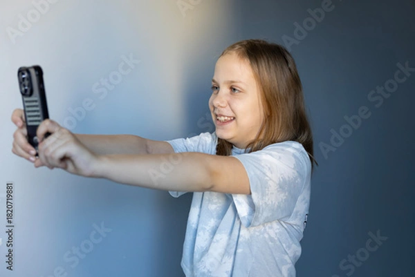 Fototapeta Child taking a selfie with a smartphone while smiling in a simple indoor setting