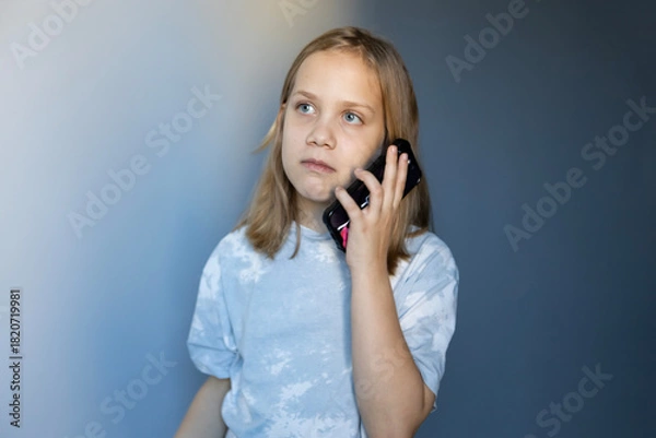 Fototapeta Young child engages in conversation on smartphone, contemplating while standing indoors in a softly lit environment