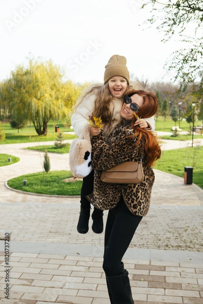 Fototapeta Mother and daughter walking hand-in-hand through a landscaped park in cool weather. Autumn vibes, warm clothes, peaceful moment.