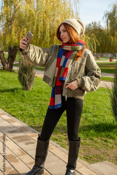 Obraz A young woman enjoying a sunny autumn or winter day outdoors, taking a cheerful selfie on a paved path in a park. Making photo.