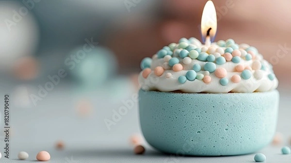 Fototapeta A single cupcake with white frosting and colorful sprinkles is topped with a lit candle. The cupcake sits on a light blue surface, with a soft, blurred backgrou