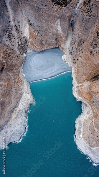 Fototapeta An aerial drone shot captures a serene, narrow lake with vibrant turquoise water, flanked by rugged, textured cliffs. A single paddleboarder navigates the calm