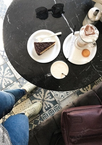 Fototapeta Cozy Coffee Break with Cake and Accessories. Top-down photo of a cozy café setting featuring a marble table with a slice of chocolate cake, a cappuccino with whipped cream and cocoa