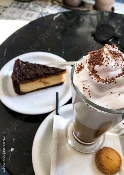 Fototapeta Creamy latte with whipped cream and chocolate shavings served with cheesecake in cozy café setting. A delicious latte topped with whipped cream and chocolate flakes served with a slice of cheesecake