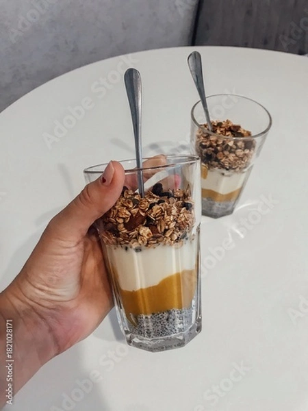 Fototapeta Hand Holding Chia Seed Parfait with Granola and Mango. A person's hand holds a glass of layered healthy dessert or breakfast, featuring chia seed pudding, a bright mango or pumpkin layer, yogurt