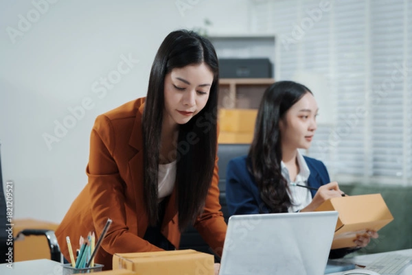 Fototapeta Asian female entrepreneurs coordinating product handover at an e-commerce office, preparing parcels for shipping logistics while collaborating professionally to manage online business operations