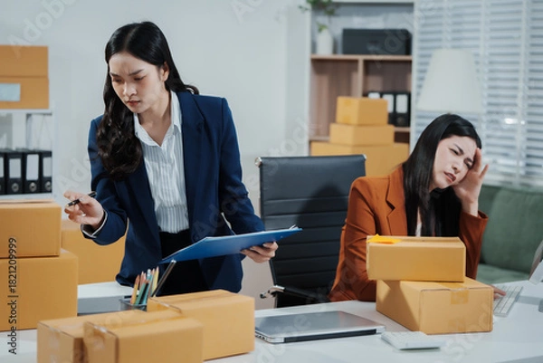 Fototapeta Two young Asian business woman stressed over poor sales, struggling with e-commerce order management, inventory, and logistics issues, showing visible frustration and pressure while handling parcels