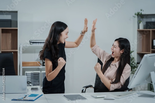 Fototapeta Two young Asian business woman celebrating success in office, reviewing results on computer screen, high-fiving, smiling, expressing achievement, digital marketing, business analysis, team finance