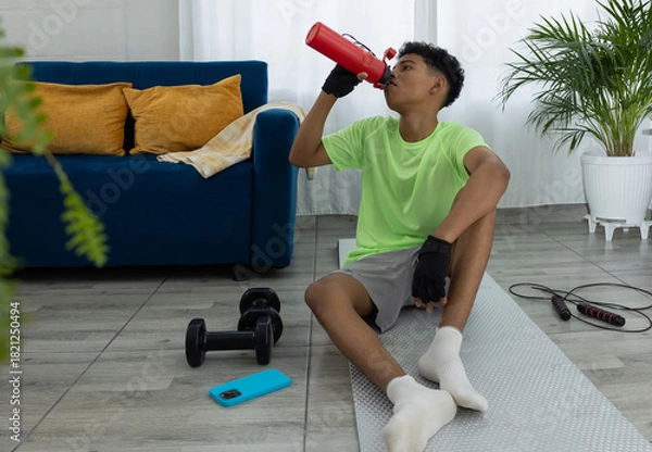 Obraz Latino teenager sitting and resting on a mat in his living room while drinking water from a red bottle, illustrating hydration and a healthy lifestyle
