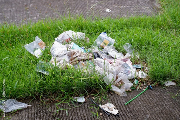 Fototapeta A pile of disposable plastic bags, bottles, and other trash sits in overgrown grass along a sidewalk. This image highlights litter, waste, and environmental neglect in urban outdoor settings.