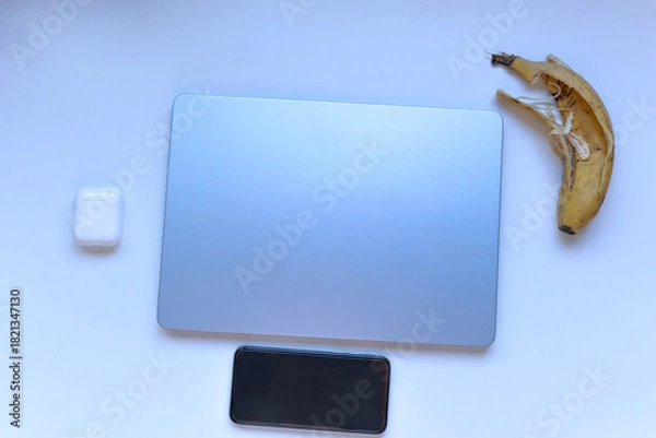 Fototapeta Minimal flat lay of a closed laptop, smartphone and wireless earbuds with a banana peel on a white background. Concept of digital detox, irony and lifestyle contrast.