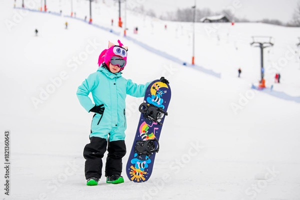 Obraz Smiling child in colorful snowboard gear standing on a snowy slope, holding a snowboard . Winter sports .